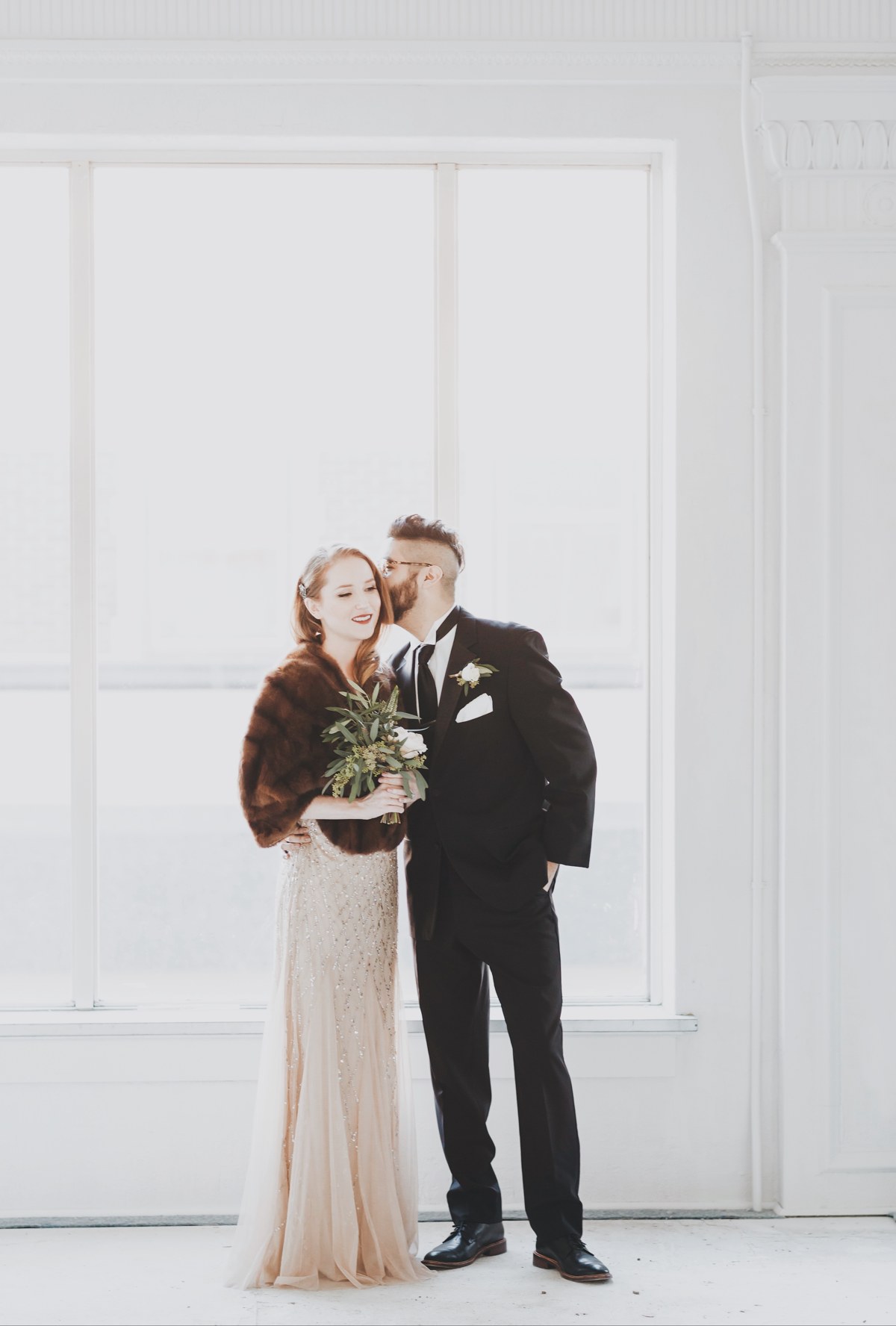 A wedding couple in soft light beside a tall white window